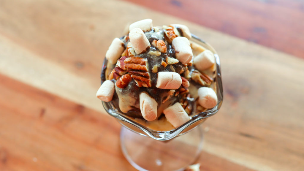 Large scoop of chocolate ice cream in a sundae cup topped with peanut butter drizzle, pecans and marshmallows