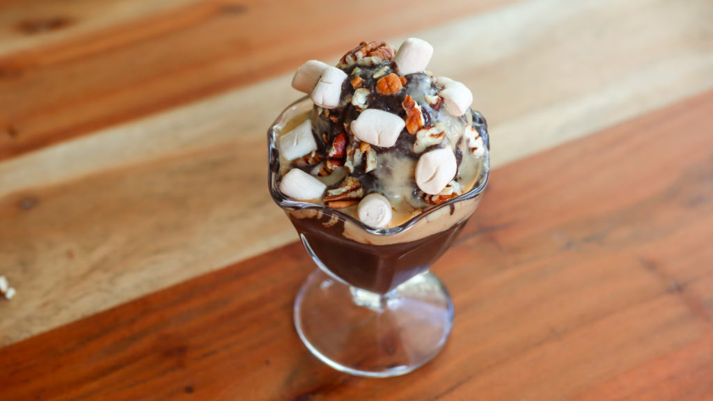 Large scoop of chocolate ice cream in a sundae cup topped with peanut butter drizzle, pecans and marshmallows