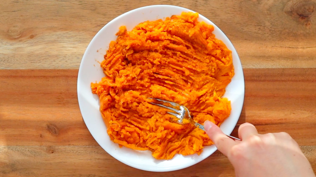 A hand holding a fork and mashing a baked sweet potato