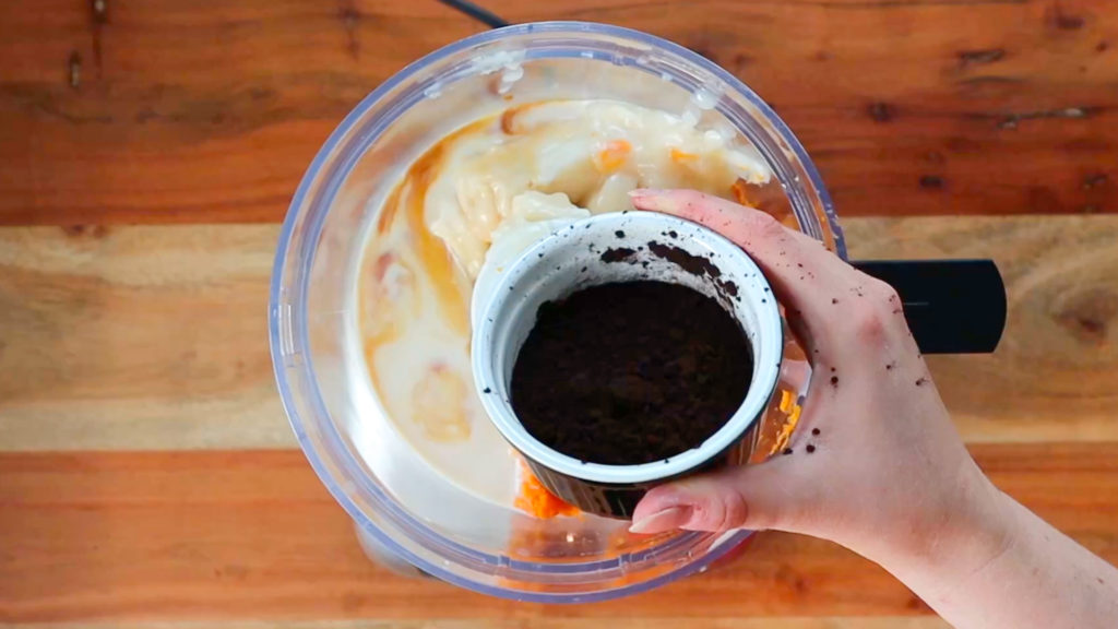 Hand holding a cup of cocoa powder, about to pour into a food processor with other ingredients
