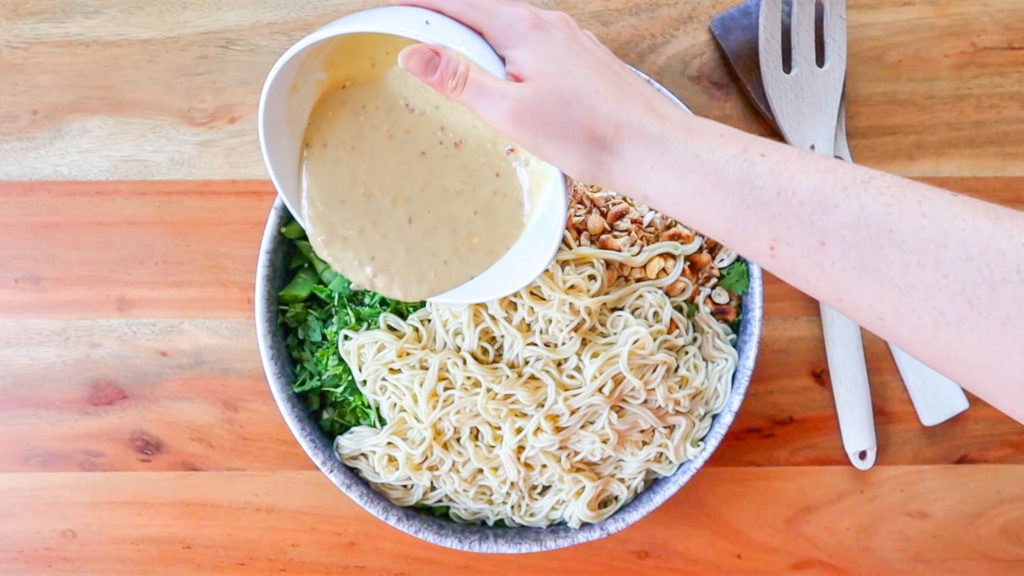 Pouring creamy dressing over a large bowl of noodle salad.