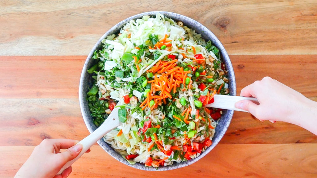 Hands holding wooden salad tossers mixing together a noodle salad in a large bowl